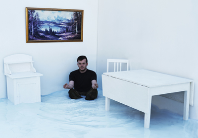 Sortkledd mann sitter i et hjørne i et hvitt rom, hvite bord og skap, naturmaleri på veggen.